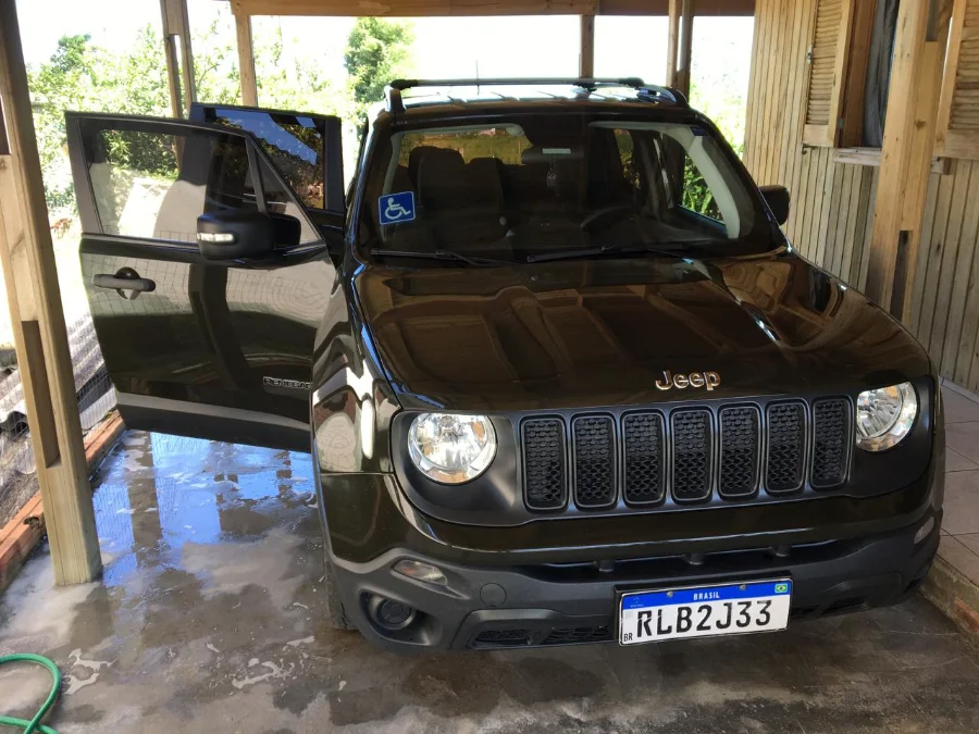 Foto frontal do Jeep Renegade verde Recon, placa RLB2J33, que foi roubado de Valdecir de Souza. O veículo está estacionado sob uma cobertura de madeira com a porta do motorista aberta e possui um adesivo azul de sinalização PCD no para-brisa dianteiro.