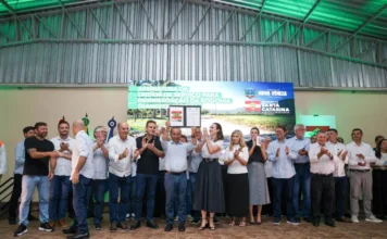 Jorginho Mello autoriza pavimentação da Transpolenteira O governador Jorginho Mello aparece ao centro da imagem, em um palco, erguendo o documento assinado da ordem de serviço para a pavimentação da Rodovia Transpolenteira. Ele está rodeado por diversas autoridades e lideranças locais, como a prefeita Angela Ghislandi, que aplaudem o ato em Nova Veneza. Ao fundo, um painel digital exibe o título do evento com os brasões oficiais do município e do Governo de Santa Catarina.