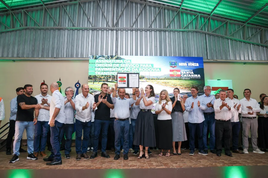 Jorginho Mello autoriza pavimentação da Transpolenteira O governador Jorginho Mello aparece ao centro da imagem, em um palco, erguendo o documento assinado da ordem de serviço para a pavimentação da Rodovia Transpolenteira. Ele está rodeado por diversas autoridades e lideranças locais, como a prefeita Angela Ghislandi, que aplaudem o ato em Nova Veneza. Ao fundo, um painel digital exibe o título do evento com os brasões oficiais do município e do Governo de Santa Catarina.
