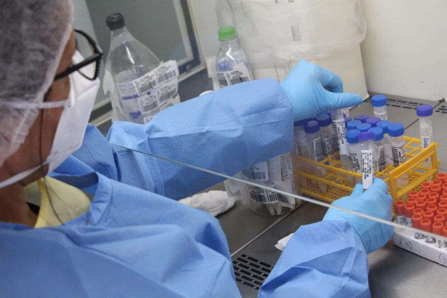 Profissional de saúde do Lacen/SC, utilizando equipamentos de proteção como luvas azuis, máscara e óculos, manuseia tubos de ensaio etiquetados em uma bancada laboratorial. A imagem detalha o trabalho técnico de análise de amostras biológicas, representando a marca de 1,1 milhão de exames atingida pelo laboratório em 2025.