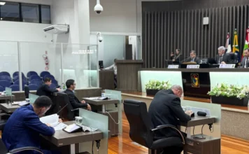 Legislativo de Araranguá retoma sessões nesta segunda Fotografia do plenário da Câmara de Vereadores de Araranguá durante uma sessão parlamentar. Em primeiro plano, vereadores vestindo terno estão sentados em suas bancadas individuais, focados na leitura de documentos e tablets. Ao fundo, os integrantes da Mesa Diretora coordenam os trabalhos atrás de uma bancada elevada, ladeada pelas bandeiras oficiais. O ambiente possui piso de madeira e iluminação clara, transmitindo o clima de trabalho legislativo.