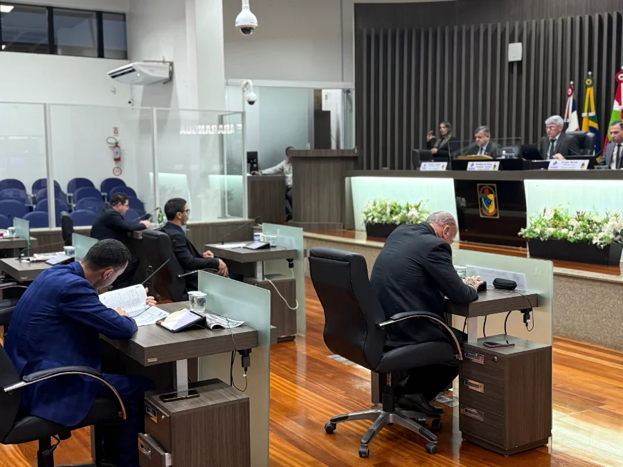 Fotografia do plenário da Câmara de Vereadores de Araranguá durante uma sessão parlamentar. Em primeiro plano, vereadores vestindo terno estão sentados em suas bancadas individuais, focados na leitura de documentos e tablets. Ao fundo, os integrantes da Mesa Diretora coordenam os trabalhos atrás de uma bancada elevada, ladeada pelas bandeiras oficiais. O ambiente possui piso de madeira e iluminação clara, transmitindo o clima de trabalho legislativo.