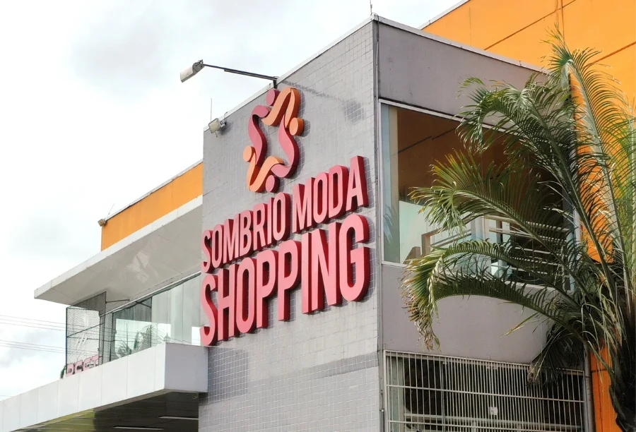 Fachada externa do Sombrio Moda Shopping, com letreiro em vermelho e logomarca, em um prédio de arquitetura moderna e palmeira na lateral direita.