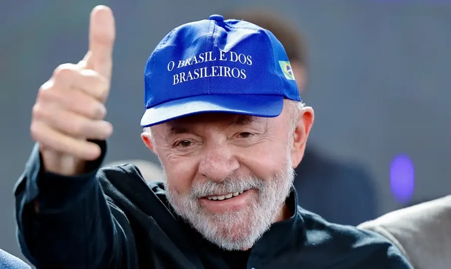 Presidente Luiz Inácio Lula da Silva sorrindo e fazendo um sinal de positivo com o polegar da mão direita. Ele veste uma camisa escura e usa um boné azul com a bandeira do Brasil na lateral e a frase 'O Brasil é dos Brasileiros' bordada em branco na frente.