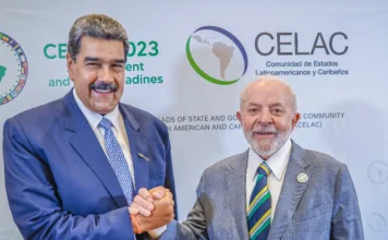 Lula condena ataque dos EUA e vê “afronta à soberania” Fotografia de plano médio do presidente da Venezuela, Nicolás Maduro (à esquerda), e do presidente do Brasil, Luiz Inácio Lula da Silva (à direita), apertando as mãos e sorrindo. Maduro veste um terno azul e Lula um terno cinza com gravata listrada nas cores verde, amarelo e azul. Ao fundo, um painel branco exibe o logotipo da CELAC. A imagem ilustra a condenação de Lula à captura de Maduro pelos EUA.