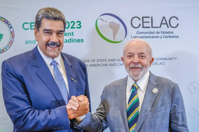 Lula condena ataque dos EUA e vê afronta à soberania Fotografia de plano médio do presidente da Venezuela, Nicolás Maduro (à esquerda), e do presidente do Brasil, Luiz Inácio Lula da Silva (à direita), apertando as mãos e sorrindo. Maduro veste um terno azul e Lula um terno cinza com gravata listrada nas cores verde, amarelo e azul. Ao fundo, um painel branco exibe o logotipo da CELAC. A imagem ilustra a condenação de Lula à captura de Maduro pelos EUA.