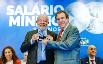 Lula critica valor do mínimo nos 90 anos da lei O presidente Luiz Inácio Lula da Silva (à esquerda) e o ministro do Trabalho e Emprego, Luiz Marinho (à direita), sorriem enquanto seguram medalhas comemorativas durante a cerimônia alusiva aos 90 anos do salário mínimo no Brasil. Atrás deles, um painel azul exibe a frase "SALÁRIO MÍNIMO 90 ANOS" e a marca do Governo Federal.