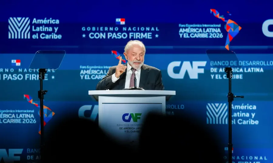 Fotografia do presidente Luiz Inácio Lula da Silva discursando em um púlpito branco com o logotipo do Banco de Desenvolvimento da América Latina (CAF). Ele veste terno escuro e gravata, gesticulando com o dedo indicador da mão direita para enfatizar seu discurso. Ao fundo, painéis azuis exibem as marcas do 