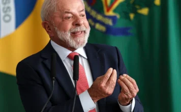 Lula diz: “Democracia será sempre uma obra em construção” Fotografia em close do presidente Luiz Inácio Lula da Silva durante discurso no Palácio do Planalto. Ele veste terno azul escuro e gravata vermelha com detalhes brancos, gesticulando com as mãos unidas em sinal de ênfase enquanto sorri. Ao fundo, aparece de forma levemente desfocada a bandeira do Brasil.