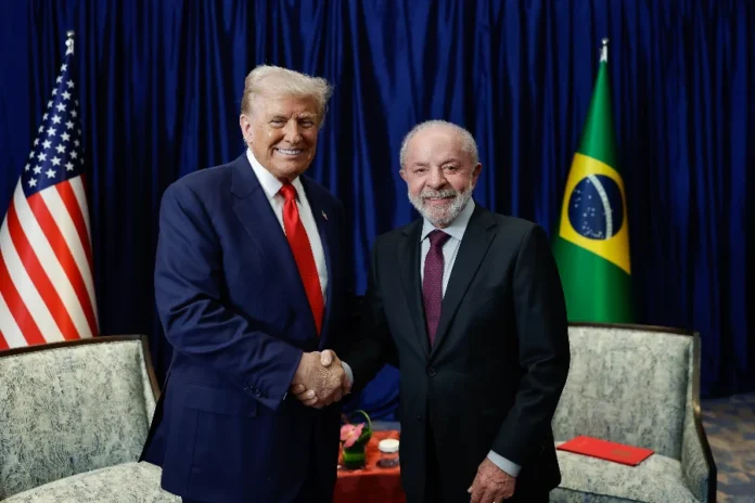 Lula e Trump se reúnem na Casa Branca em março O presidente Donald Trump (esquerda) e o presidente Luiz Inácio Lula da Silva (direita) apertam as mãos e sorriem para a câmera, posicionados em frente a um fundo de cortina azul com as bandeiras dos Estados Unidos e do Brasil nas laterais. A imagem ilustra a confirmação feita por Lula de um futuro encontro bilateral na Casa Branca em março para discutir as relações entre as duas nações.