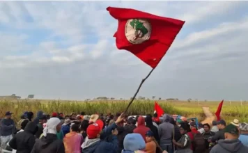 Lula encerra encontro nacional do MST nesta sexta na Bahia Grupo de camponeses do Movimento dos Trabalhadores Rurais Sem Terra (MST) reunidos em área rural, observando uma grande bandeira vermelha do movimento hasteada improvisadamente, simbolizando a mobilização pela reforma agrária.