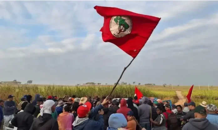 Lula encerra encontro nacional do MST nesta sexta na Bahia Grupo de camponeses do Movimento dos Trabalhadores Rurais Sem Terra (MST) reunidos em área rural, observando uma grande bandeira vermelha do movimento hasteada improvisadamente, simbolizando a mobilização pela reforma agrária.