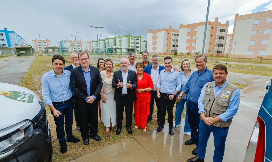 O presidente Luiz Inácio Lula da Silva, sorrindo e fazendo sinal de positivo com os polegares, posa para foto cercado por autoridades e representantes em frente aos novos prédios de apartamentos do conjunto habitacional entregue pelo programa Minha Casa, Minha Vida em Rio Grande, no Rio Grande do Sul. O céu está nublado e o chão molhado, indicando chuva recente.