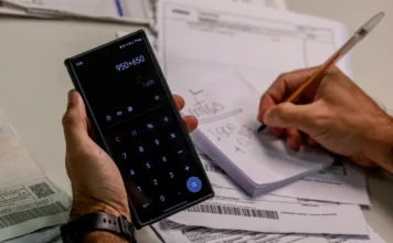 Lula sanciona lei do Código de Defesa do Contribuinte Mãos de uma pessoa calculando valores na calculadora do celular e fazendo anotações com uma caneta em um bloco de papel. Vários boletos, contas e documentos estão espalhados sobre uma superfície branca ao redor.