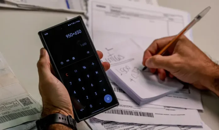 Lula sanciona lei do Código de Defesa do Contribuinte Mãos de uma pessoa calculando valores na calculadora do celular e fazendo anotações com uma caneta em um bloco de papel. Vários boletos, contas e documentos estão espalhados sobre uma superfície branca ao redor.