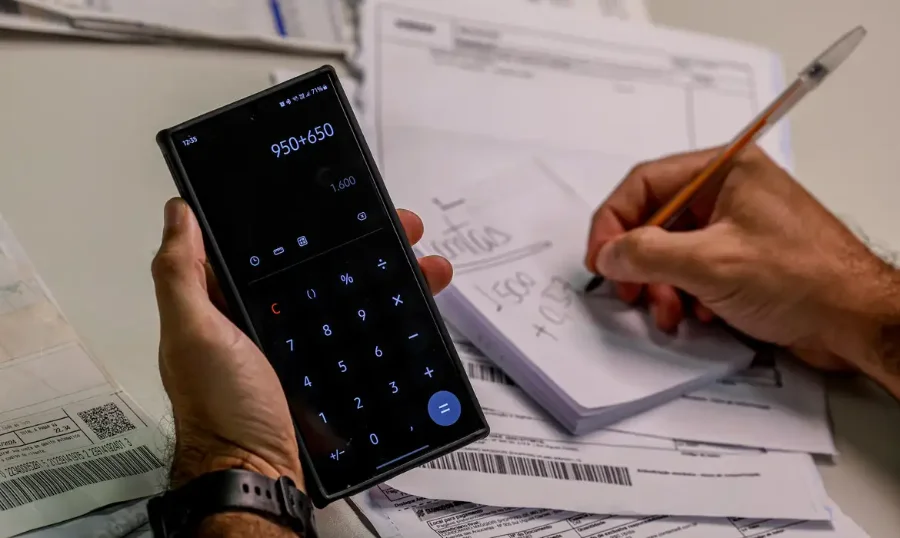 Lula sanciona lei do Código de Defesa do Contribuinte Mãos de uma pessoa calculando valores na calculadora do celular e fazendo anotações com uma caneta em um bloco de papel. Vários boletos, contas e documentos estão espalhados sobre uma superfície branca ao redor.