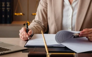 MPSC abre inquérito por falhas em escolas de Sombrio Mãos de uma pessoa em traje profissional assinando e revisando documentos sobre uma mesa de escritório. Ao fundo, uma balança da justiça e livros jurídicos simbolizam a atuação legal e a abertura do inquérito civil pelo MPSC em Sombrio.