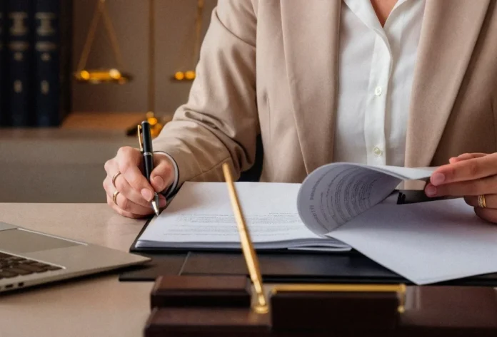 MPSC abre inquérito por falhas em escolas de Sombrio Mãos de uma pessoa em traje profissional assinando e revisando documentos sobre uma mesa de escritório. Ao fundo, uma balança da justiça e livros jurídicos simbolizam a atuação legal e a abertura do inquérito civil pelo MPSC em Sombrio.