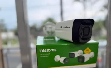Maracajá amplia monitoramento em escolas e órgãos públicos Fotografia em primeiro plano de uma câmera de segurança do tipo "bullet" da marca Intelbras, posicionada sobre sua caixa original de cor verde e branca. Uma mão segura o equipamento em um ambiente externo com fundo levemente desfocado. A imagem ilustra os novos dispositivos tecnológicos adquiridos pelo município de Maracajá para ampliar o monitoramento de segurança.
