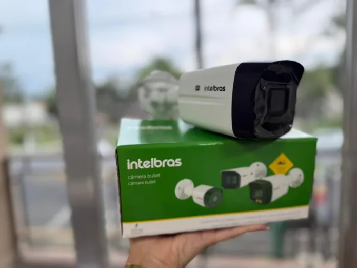 Maracajá amplia monitoramento em escolas e órgãos públicos Fotografia em primeiro plano de uma câmera de segurança do tipo "bullet" da marca Intelbras, posicionada sobre sua caixa original de cor verde e branca. Uma mão segura o equipamento em um ambiente externo com fundo levemente desfocado. A imagem ilustra os novos dispositivos tecnológicos adquiridos pelo município de Maracajá para ampliar o monitoramento de segurança.