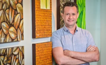 Marcus Lacerda assume direção de programa global da OMS Foto de Marcus Vinicius Guimarães Lacerda, médico infectologista da Fiocruz e novo diretor do Programa Especial para Pesquisa e Treinamento em Doenças Tropicais (TDR) da OMS. Ele é um homem de meia-idade, sorrindo, com os braços cruzados e vestindo uma camisa cinza, encostado em uma parede decorada com quadros coloridos.