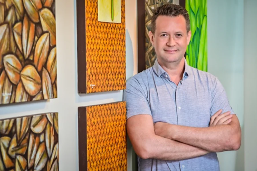 Foto de Marcus Vinicius Guimarães Lacerda, médico infectologista da Fiocruz e novo diretor do Programa Especial para Pesquisa e Treinamento em Doenças Tropicais (TDR) da OMS. Ele é um homem de meia-idade, sorrindo, com os braços cruzados e vestindo uma camisa cinza, encostado em uma parede decorada com quadros coloridos.