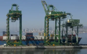 Mercosul-UE: entidades celebram acordo histórico Grandes guindastes verdes em um porto movimentado com contêineres e um navio, com a cidade ao fundo.