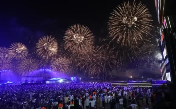 Metas de 2026: Médicos dão dicas para não desistir em janeiro Fotografia noturna de uma grande multidão de costas, observando um espetáculo de fogo-de-artifício dourado e brilhante que preenche quase todo o céu. No centro, ao fundo, vê-se um palco iluminado com luzes azuis e ecrãs, indicando um concerto ou evento de celebração. Muitas pessoas na audiência seguram telemóveis para registar o momento. A imagem ilustra as celebrações de Ano Novo e a magnitude do evento cultural.