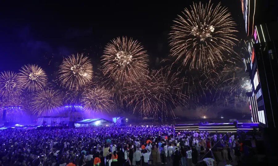Fotografia noturna de uma grande multidão de costas, observando um espetáculo de fogo-de-artifício dourado e brilhante que preenche quase todo o céu. No centro, ao fundo, vê-se um palco iluminado com luzes azuis e ecrãs, indicando um concerto ou evento de celebração. Muitas pessoas na audiência seguram telemóveis para registar o momento. A imagem ilustra as celebrações de Ano Novo e a magnitude do evento cultural.