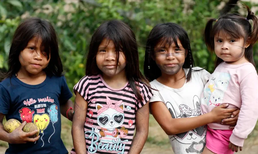 Quatro meninas indígenas pequenas, com cabelos escuros e camisetas estampadas, posam juntas olhando para a câmera em uma área com vegetação ao fundo.