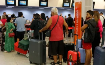 Ministério define regras para bagagens vindas do exterior Fila de passageiros com malas aguardando atendimento nos balcões de check-in da companhia aérea GOL em um aeroporto. Monitores acima dos balcões mostram voos para Miami e Orlando. Algumas pessoas na fila usam máscaras de proteção facial. Um homem de regata vermelha e shorts pretos está em destaque no centro da fila, segurando uma mala cinza.
