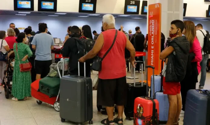 Ministério define regras para bagagens vindas do exterior (1) Fila de passageiros com malas aguardando atendimento nos balcões de check-in da companhia aérea GOL em um aeroporto. Monitores acima dos balcões mostram voos para Miami e Orlando. Algumas pessoas na fila usam máscaras de proteção facial. Um homem de regata vermelha e shorts pretos está em destaque no centro da fila, segurando uma mala cinza.