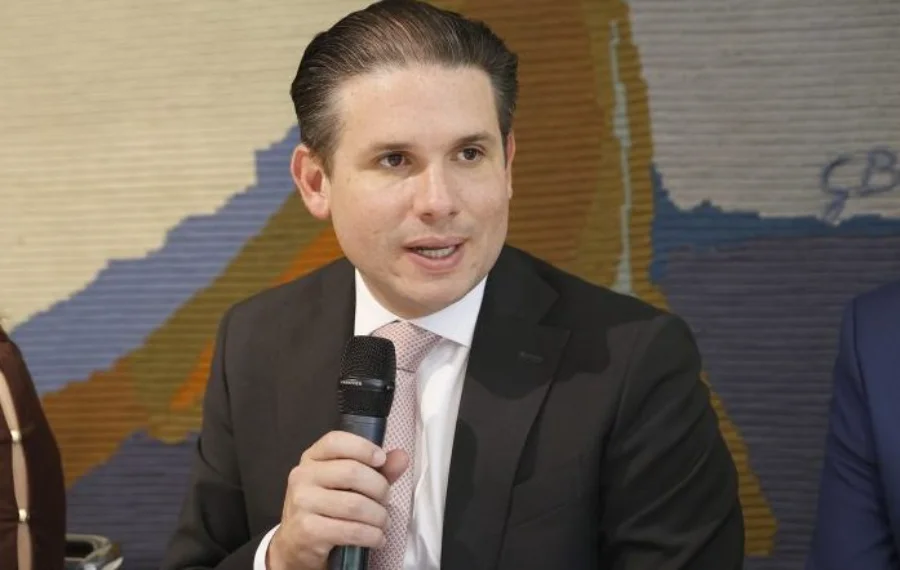 Fotografia em plano médio do presidente da Câmara, Hugo Motta. Ele é um homem jovem, de pele clara, cabelos escuros penteados para trás e veste terno escuro com gravata clara. Ele segura um microfone preto próximo à boca, com uma expressão de fala séria e focada. Ao fundo, uma parede com textura em tons terrosos e azulados.