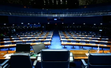 Mudanças partidárias alteram forças no Senado em 2026 Visão geral do plenário do Senado Federal vazio, com suas fileiras de assentos azuis e mesas de madeira, a partir da perspectiva da Mesa Diretora. A imagem ilustra o local onde ocorrem as disputas políticas e a nova correlação de forças entre as bancadas partidárias para o ano legislativo de 2026, conforme detalhado na matéria.