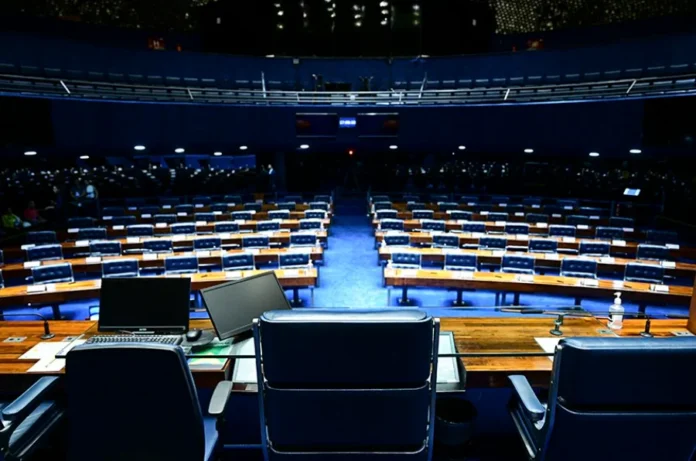 Mudanças partidárias alteram forças no Senado em 2026 Visão geral do plenário do Senado Federal vazio, com suas fileiras de assentos azuis e mesas de madeira, a partir da perspectiva da Mesa Diretora. A imagem ilustra o local onde ocorrem as disputas políticas e a nova correlação de forças entre as bancadas partidárias para o ano legislativo de 2026, conforme detalhado na matéria.