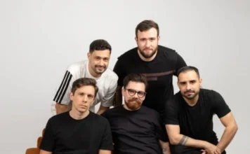 Nova Cidade lança músicas gravadas com mentor americano Os cinco integrantes da banda Nova Cidade posam para uma foto promocional em um estúdio com fundo branco. Eles estão agrupados, vestindo camisetas casuais nas cores preto e branco, e olham diretamente para a câmera. A imagem transmite a identidade visual do grupo para o lançamento de seu novo projeto musical.