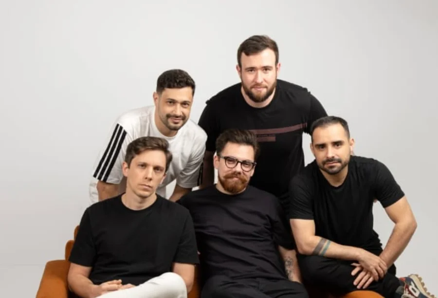 Nova Cidade lança músicas gravadas com mentor americano Os cinco integrantes da banda Nova Cidade posam para uma foto promocional em um estúdio com fundo branco. Eles estão agrupados, vestindo camisetas casuais nas cores preto e branco, e olham diretamente para a câmera. A imagem transmite a identidade visual do grupo para o lançamento de seu novo projeto musical.