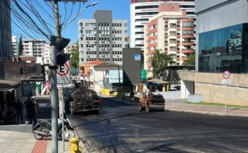 Obras fecham trecho da Coronel Pedro Benedet em Criciúma Fotografia urbana da Rua Coronel Pedro Benedet, no Centro de Criciúma, mostrando o trabalho de recapeamento asfáltico. No centro da imagem, um rolo compactador e um caminhão trabalham sobre a pista coberta por asfalto novo e escuro. Ao fundo, prédios residenciais e comerciais sob um céu azul. À esquerda, uma calçada com hidrante amarelo e pedestres observando a obra.