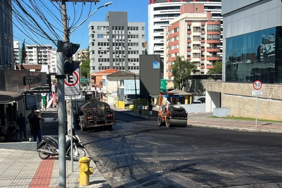 Obras fecham trecho da Coronel Pedro Benedet em Criciúma Fotografia urbana da Rua Coronel Pedro Benedet, no Centro de Criciúma, mostrando o trabalho de recapeamento asfáltico. No centro da imagem, um rolo compactador e um caminhão trabalham sobre a pista coberta por asfalto novo e escuro. Ao fundo, prédios residenciais e comerciais sob um céu azul. À esquerda, uma calçada com hidrante amarelo e pedestres observando a obra.
