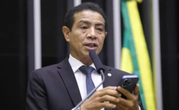 PL quer proibir imagens religiosas em desfiles de Carnaval Fotografia do deputado Pastor Gil (PL-MA), autor do Projeto de Lei 830/25. Ele é um homem pardo, de cabelos curtos e escuros, veste terno preto, camisa branca e gravata azul. Na imagem, ele aparece falando ao microfone em um púlpito, enquanto segura um smartphone com as duas mãos. O fundo está levemente desfocado, sugerindo o ambiente da Câmara dos Deputados.