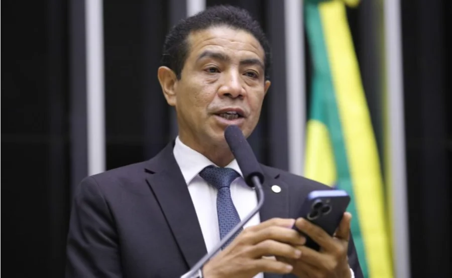 Fotografia do deputado Pastor Gil (PL-MA), autor do Projeto de Lei 830/25. Ele é um homem pardo, de cabelos curtos e escuros, veste terno preto, camisa branca e gravata azul. Na imagem, ele aparece falando ao microfone em um púlpito, enquanto segura um smartphone com as duas mãos. O fundo está levemente desfocado, sugerindo o ambiente da Câmara dos Deputados.