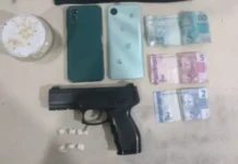PM prende dupla por tráfico de drogas em Praia Grande Fotografia de materiais apreendidos pela Polícia Militar sobre uma superfície clara. A imagem exibe um simulacro de pistola preta, sete pequenas embalagens plásticas brancas (petecas) contendo substância análoga à cocaína, dois smartphones (um verde escuro e um verde claro) e três cédulas de Real (uma de 100, uma de 5 e uma de 2), totalizando 107 reais.