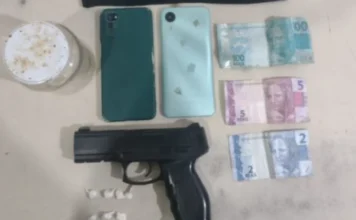 PM prende dupla por tráfico de drogas em Praia Grande Fotografia de materiais apreendidos pela Polícia Militar sobre uma superfície clara. A imagem exibe um simulacro de pistola preta, sete pequenas embalagens plásticas brancas (petecas) contendo substância análoga à cocaína, dois smartphones (um verde escuro e um verde claro) e três cédulas de Real (uma de 100, uma de 5 e uma de 2), totalizando 107 reais.