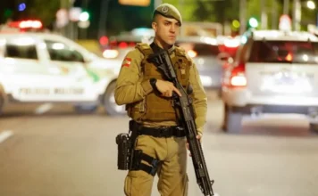 PMSC 190 anos: SC consolida título de estado mais seguro Policial militar fardado segurando uma espingarda tática em uma rua à noite. Ele usa boina verde e colete balístico; ao fundo, luzes de viaturas desfocadas iluminam a cena. Ilustra a consolidação de SC como o estado mais seguro do país nos 190 anos da PMSC.