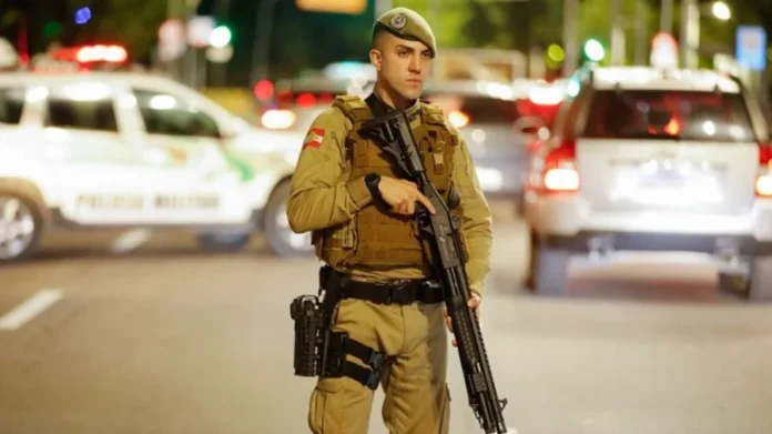 PMSC 190 anos SC consolida título de estado mais seguro Policial militar fardado segurando uma espingarda tática em uma rua à noite. Ele usa boina verde e colete balístico; ao fundo, luzes de viaturas desfocadas iluminam a cena. Ilustra a consolidação de SC como o estado mais seguro do país nos 190 anos da PMSC.