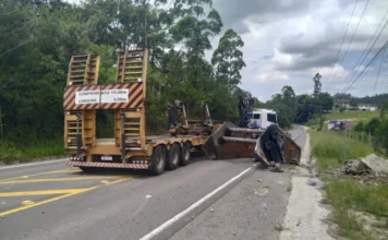 Peça de metal cai de caminhão e trava via em Siderópolis Foto mostra uma grande peça metálica caída de um caminhão prancha amarelo, bloqueando parcialmente uma das faixas da Rua Lino de Araújo, em Siderópolis. A estrutura escura está apoiada entre o asfalto e o acostamento, enquanto o caminhão permanece parado logo à frente. Ao fundo, é possível ver uma área de vegetação e a continuidade da via.
