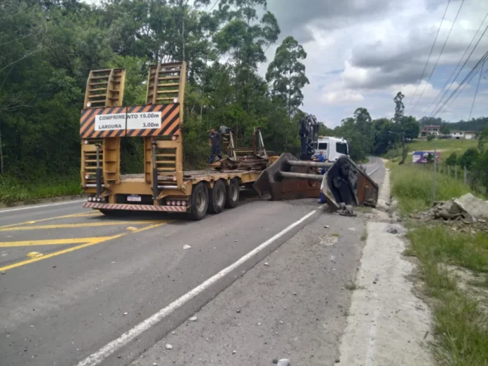 Peça de metal cai de caminhão e trava via em Siderópolis Foto mostra uma grande peça metálica caída de um caminhão prancha amarelo, bloqueando parcialmente uma das faixas da Rua Lino de Araújo, em Siderópolis. A estrutura escura está apoiada entre o asfalto e o acostamento, enquanto o caminhão permanece parado logo à frente. Ao fundo, é possível ver uma área de vegetação e a continuidade da via.