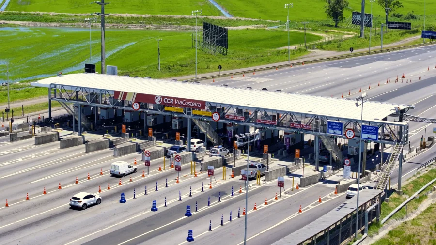 Pedágio na BR-101 saiba como evitar filas no verão Vista aérea de uma praça de pedágio da ViaCosteira na BR-101 Sul, sob luz do dia, mostrando diversas pistas com fluxo de veículos. Pistas exclusivas para cobrança automática estão sinalizadas, ilustrando as alternativas para evitar filas durante viagens de verão.