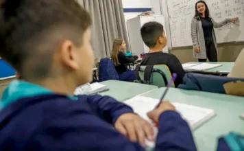 Piso do magistério vai a R$ 5.130 em 2026 com ganho real Uma foto em ângulo baixo captura uma sala de aula ativa. Em primeiro plano, ligeiramente desfocado, um aluno de casaco azul escreve em seu caderno com um lápis roxo. Mais atrás, uma aluna de preto olha atentamente para a frente. Ao fundo, uma professora sorridente, vestindo um casaco cinza, está em pé ao lado de um quadro branco preenchido com equações matemáticas e anotações, apontando para o conteúdo. A sala é iluminada e organizada, com carteiras e armários visíveis.