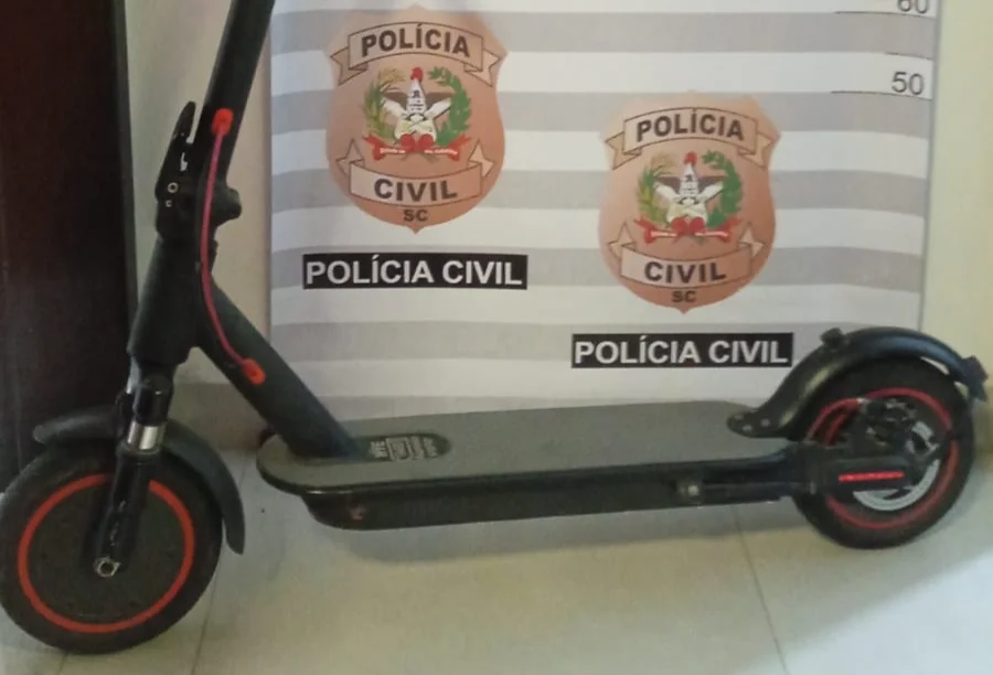 Patinete elétrico preto com detalhes em vermelho, modelo HYE, posicionado em frente a um painel oficial da Polícia Civil de Santa Catarina com marcações de altura. O equipamento foi recuperado em Passo de Torres após ser desviado de Balneário Camboriú.